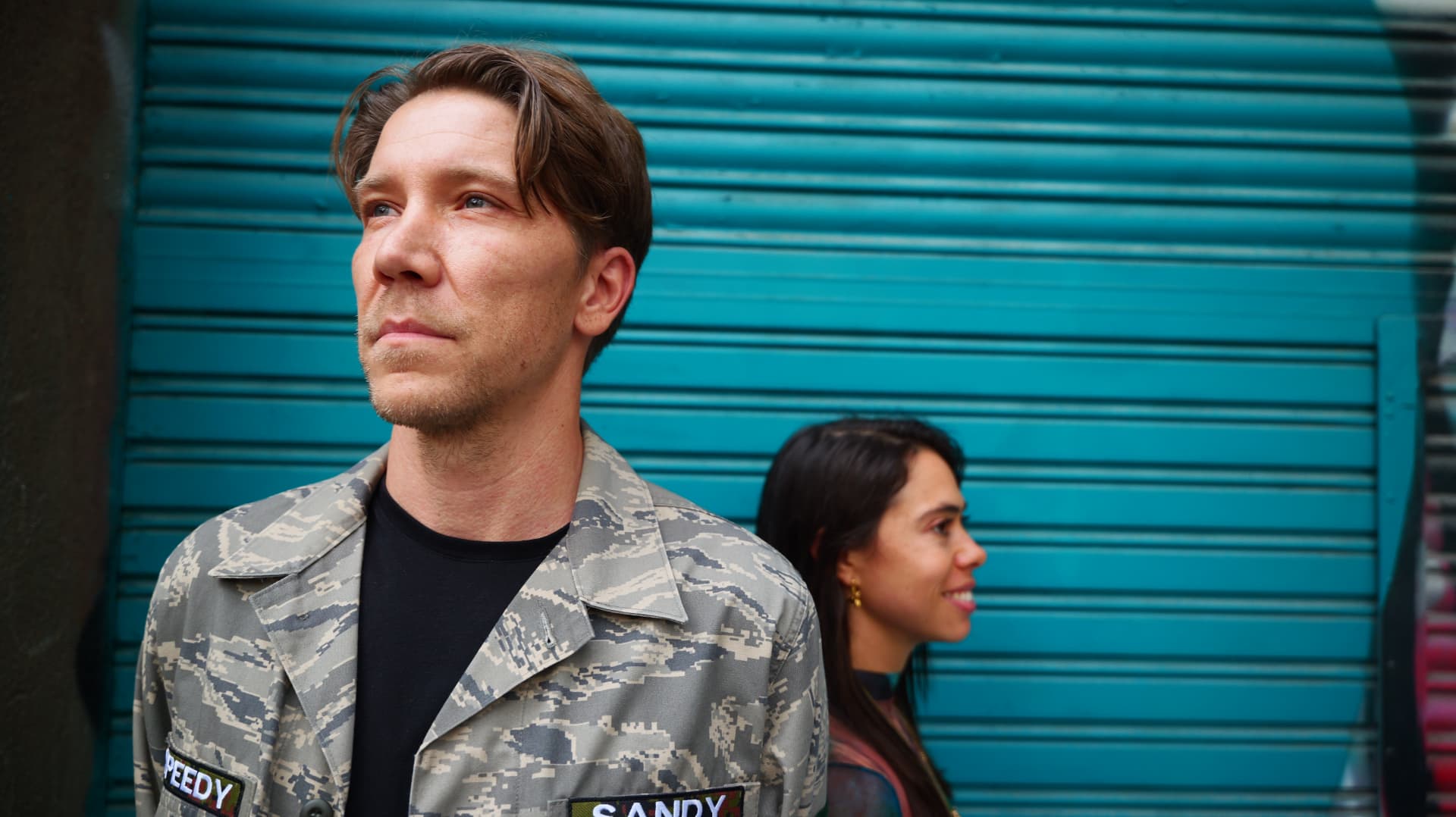 Man in a camouflage jacket and a woman smiling in front of a teal shutter
