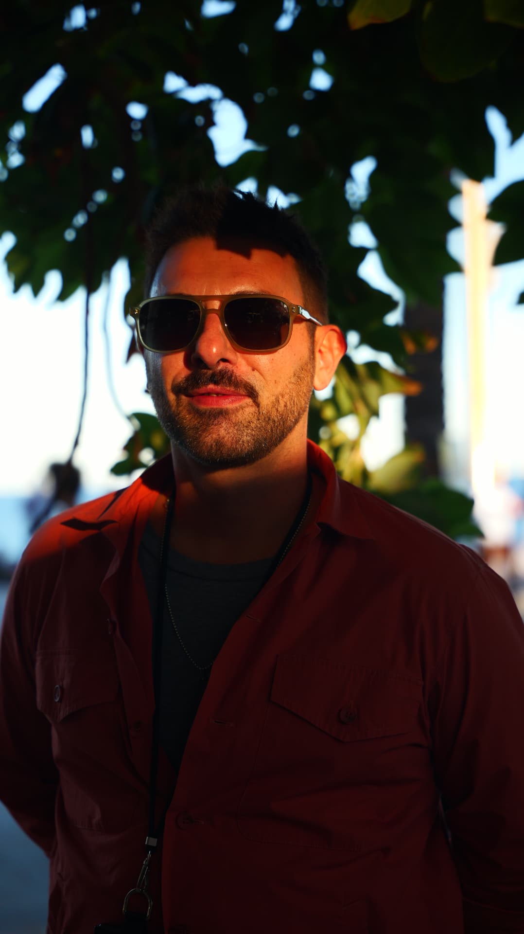 Man in sunglasses and a red shirt under tree shade at sunset