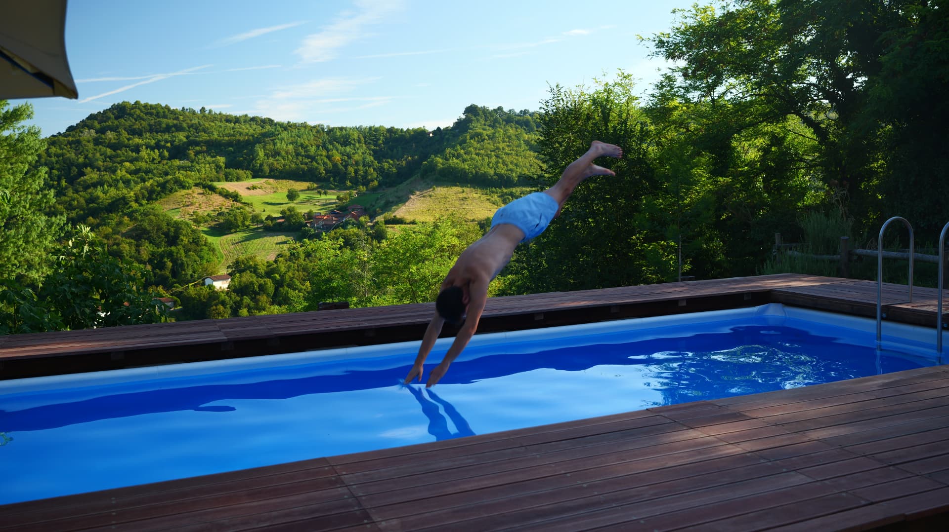 Man diving into a blue hillside pool overlooking a green Italian valley