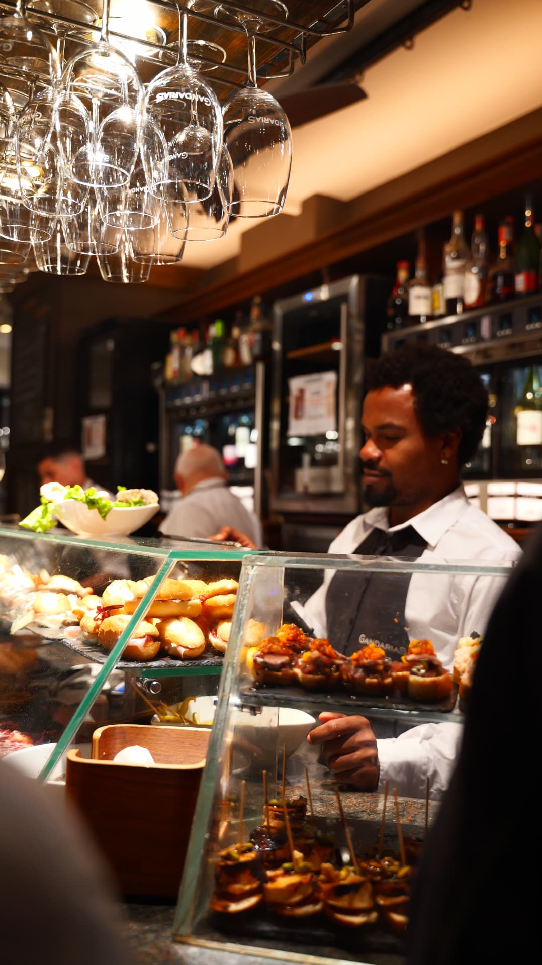 Bartender smiling behind a counter of pintxos in a warmly lit San Sebastián bar