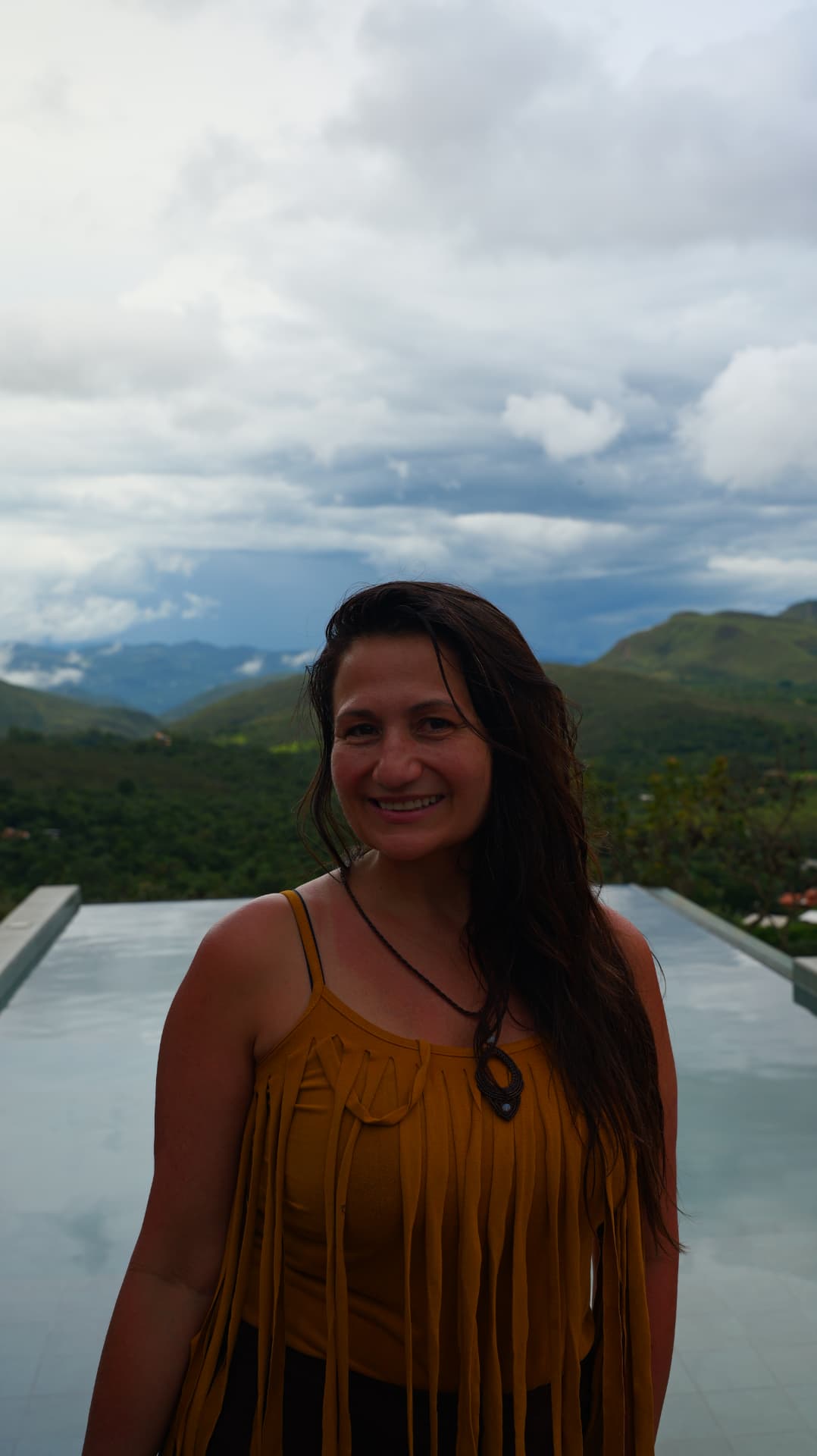 Smiling woman in a mustard fringed top with mountains and an infinity pool behind her