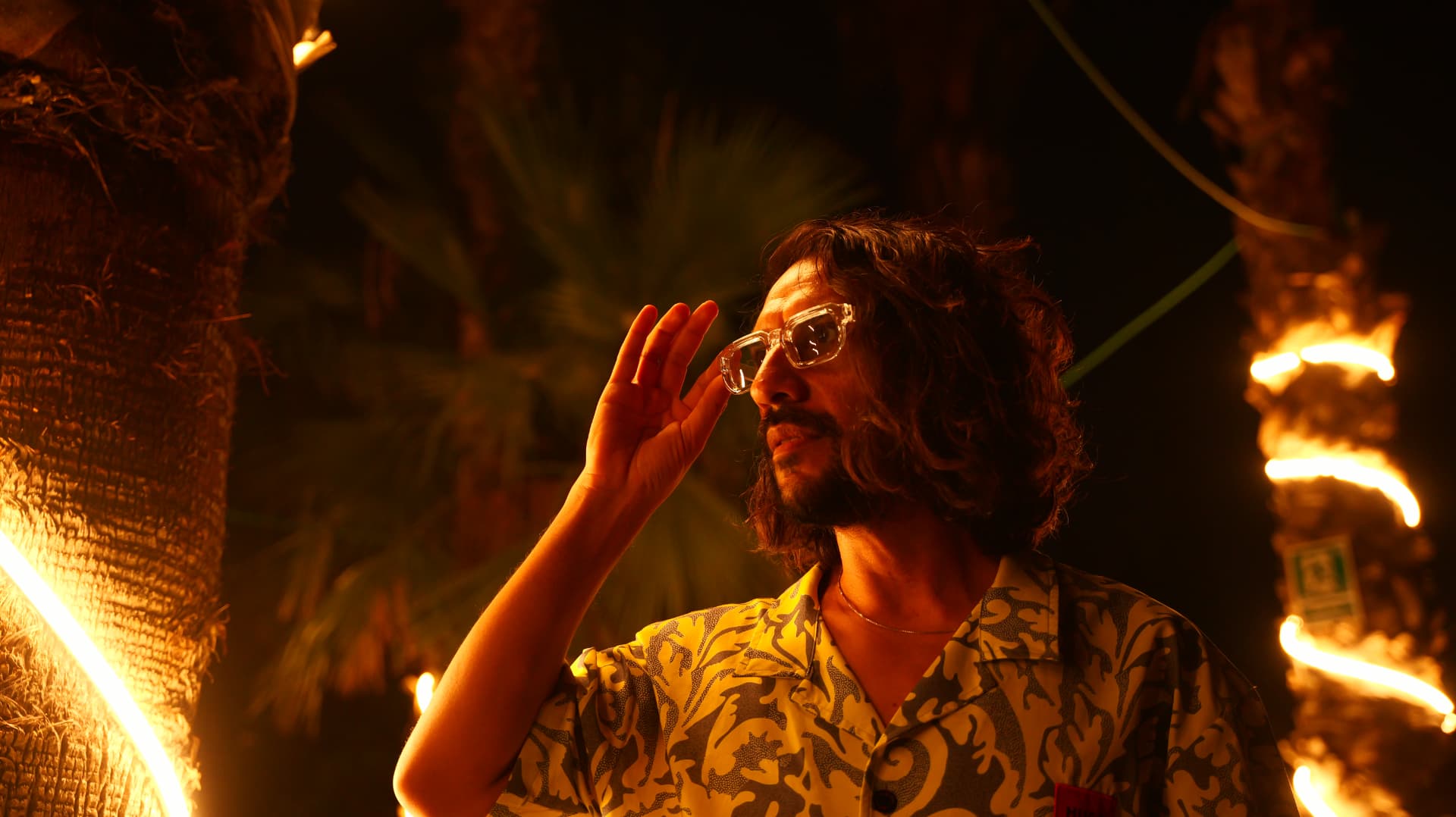 Man adjusting clear glasses beside palm torches in warm firelight