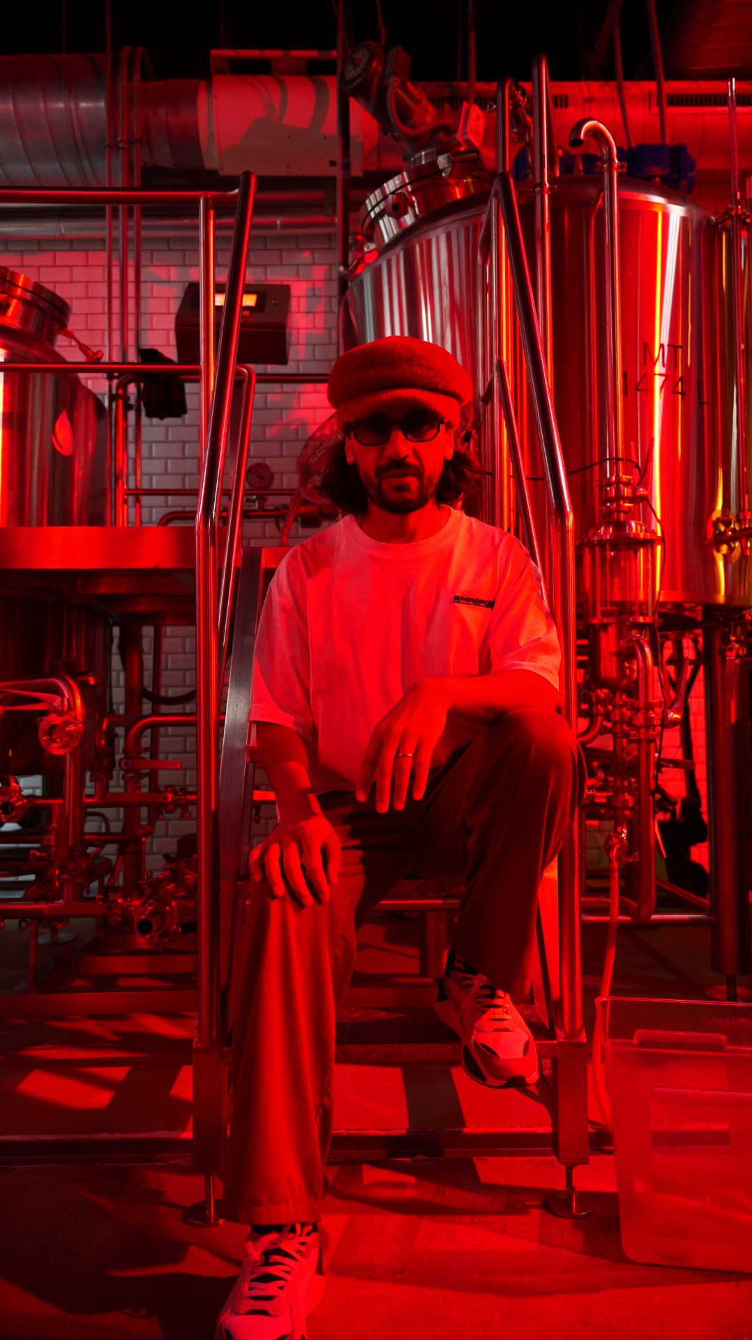 Man in a beret seated among brewery tanks in vivid red light