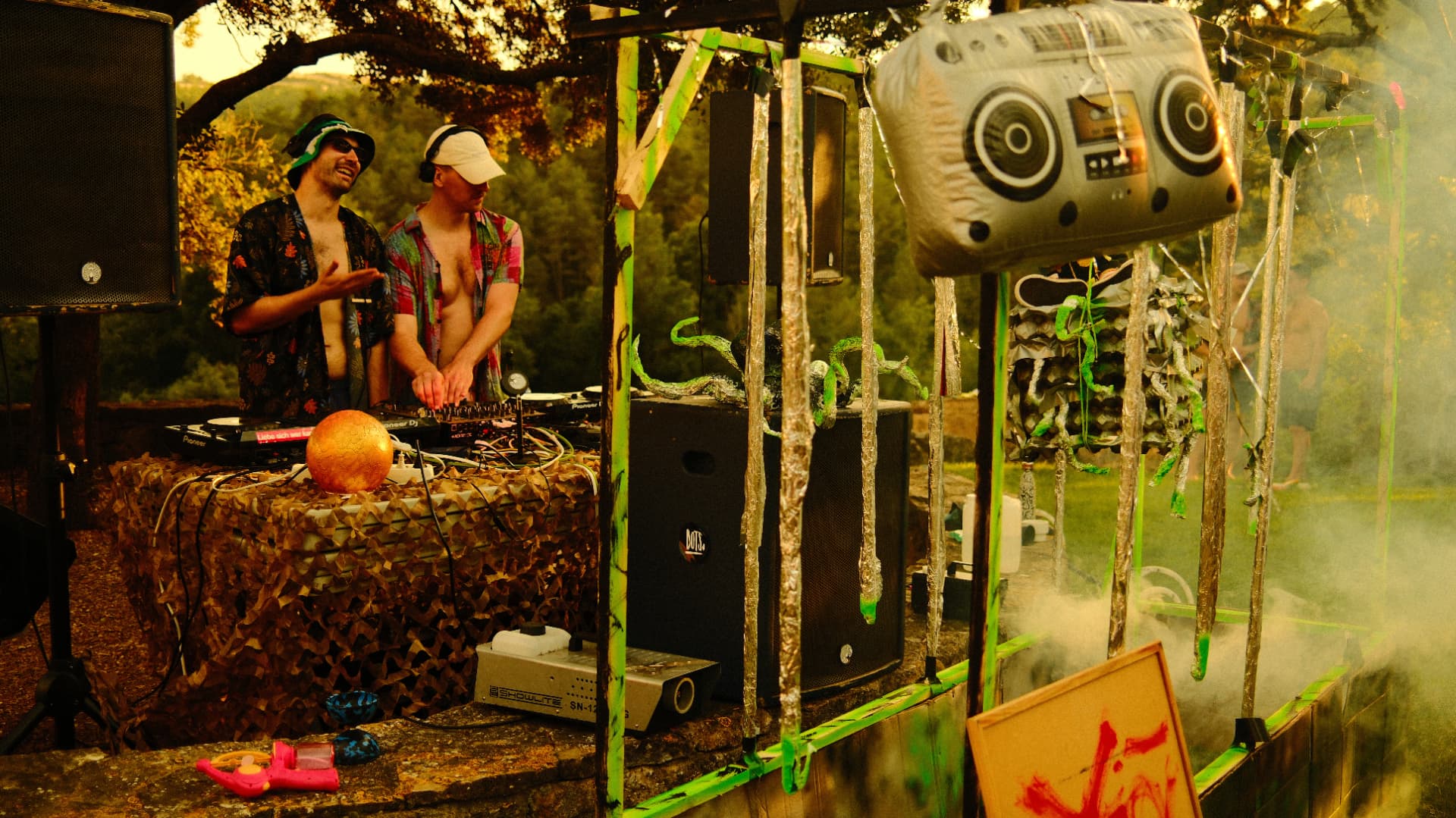Two DJs playing at a decorated outdoor booth in golden evening haze
