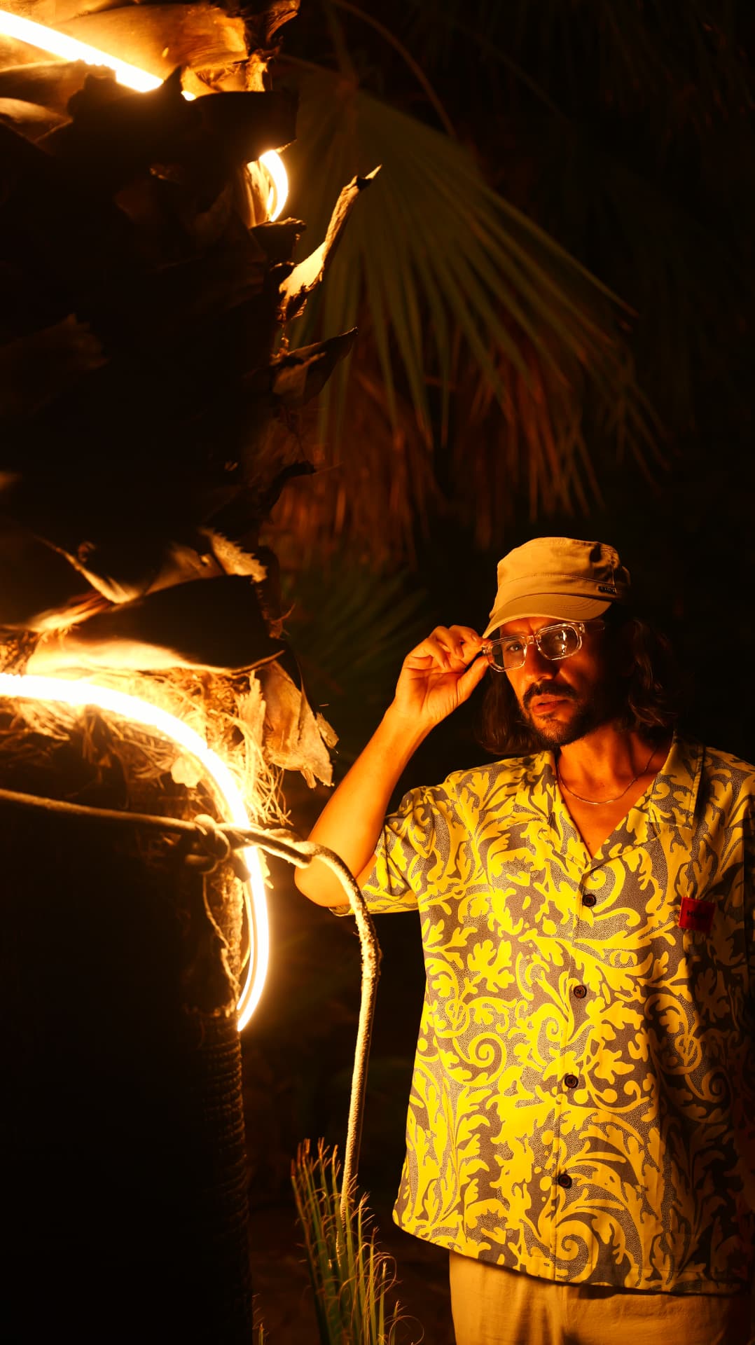 Man in a cap beside glowing neon light rings on a palm tree at night