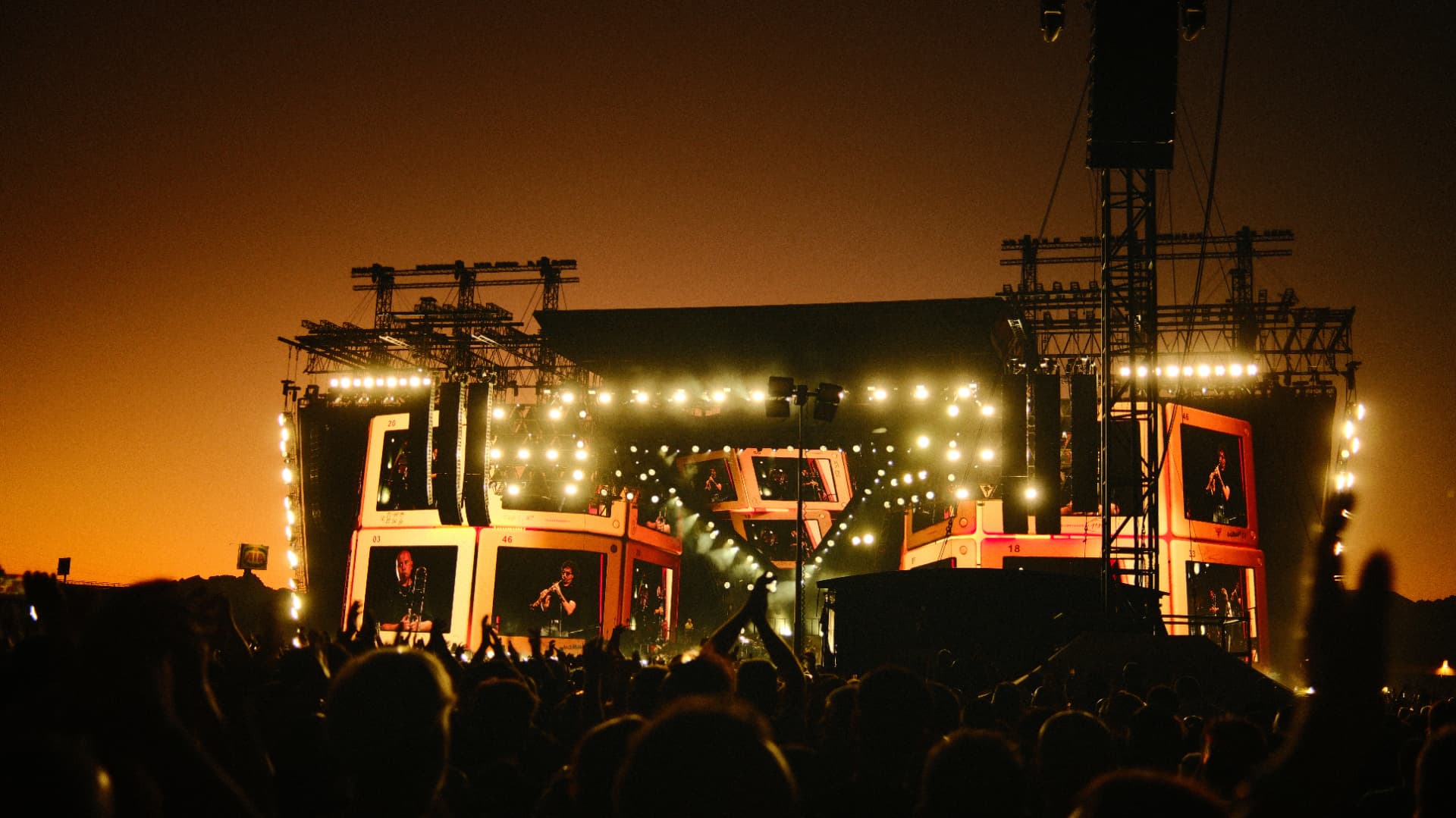 Festival crowd in front of a brightly lit stage under an orange night sky