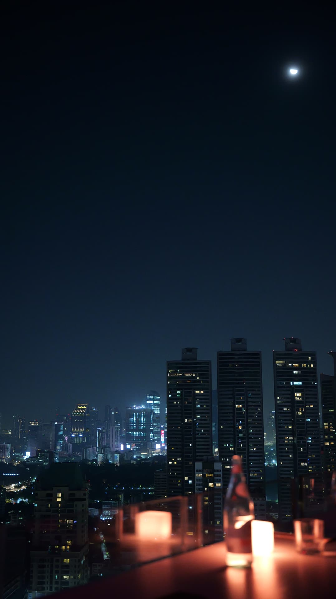 Bangkok skyline beneath the moon, viewed from a candlelit rooftop table