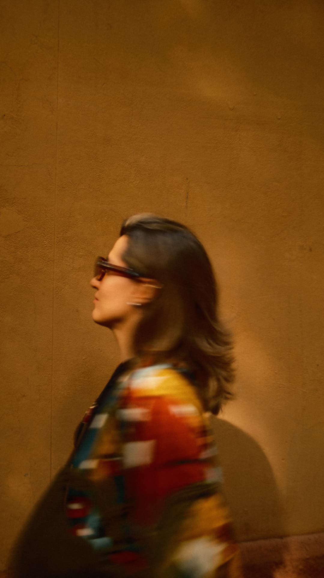 Motion-blurred profile of a woman in sunglasses against a warm ochre wall