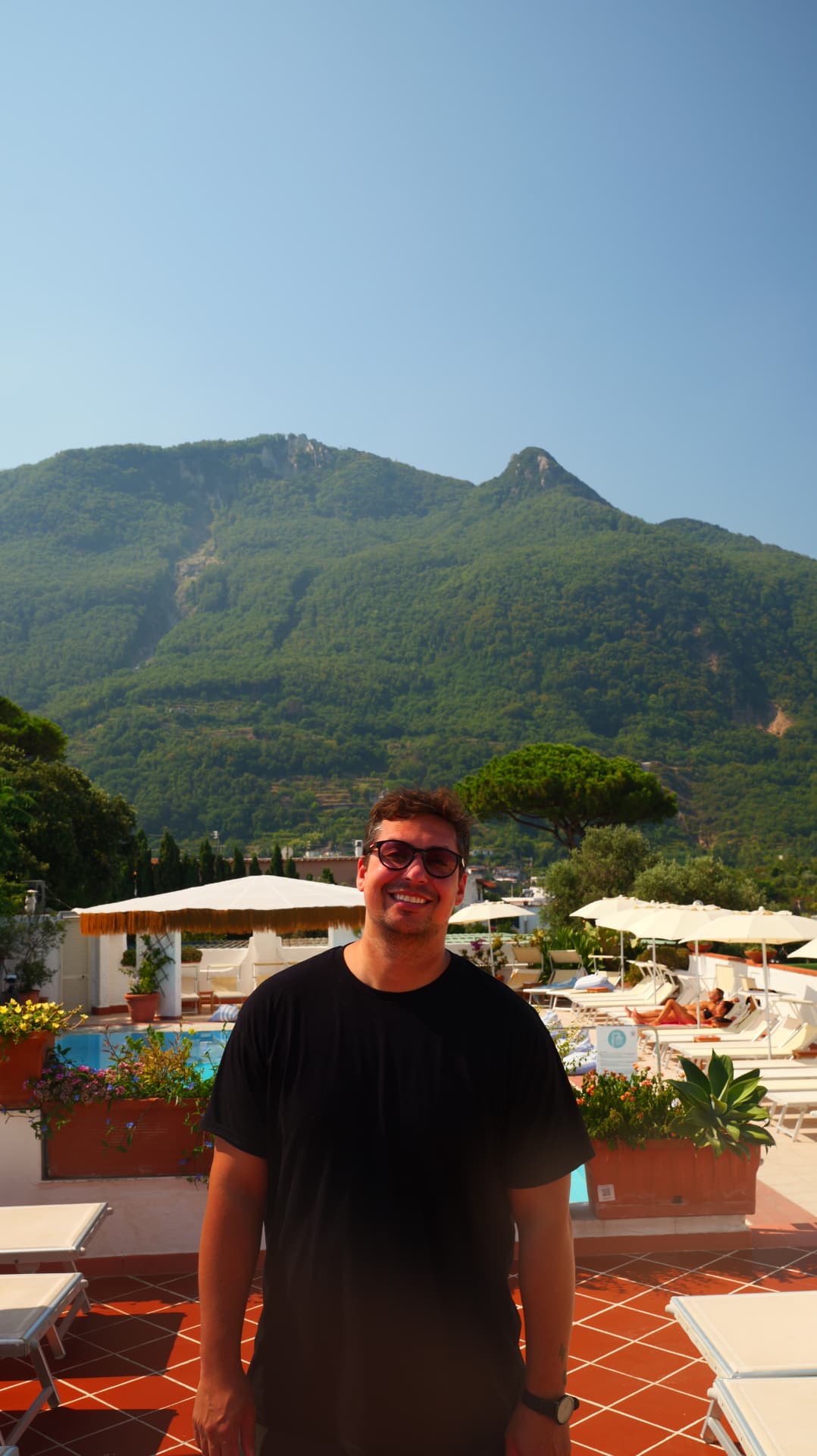 Smiling man in sunglasses on a pool terrace with a green mountain behind