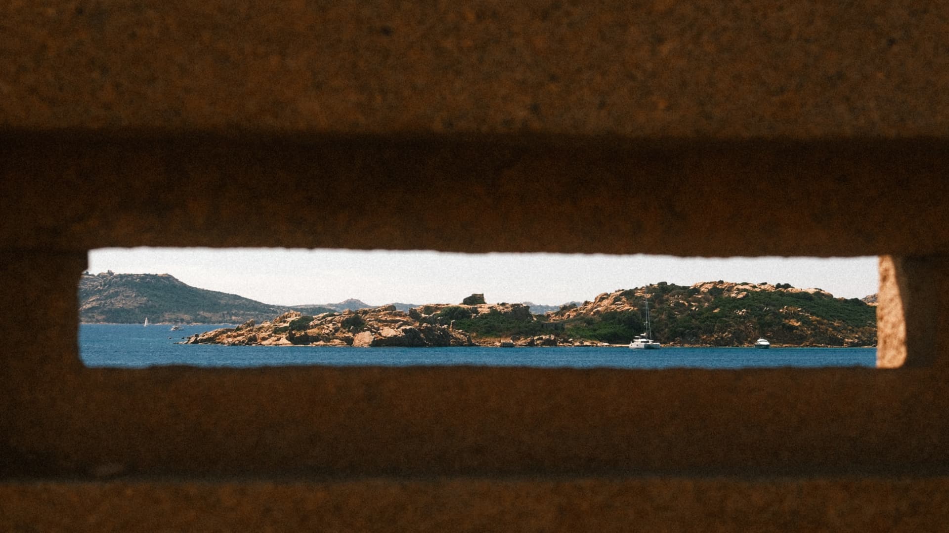 Sardinian coastline and sailing boats seen through a narrow stone slit