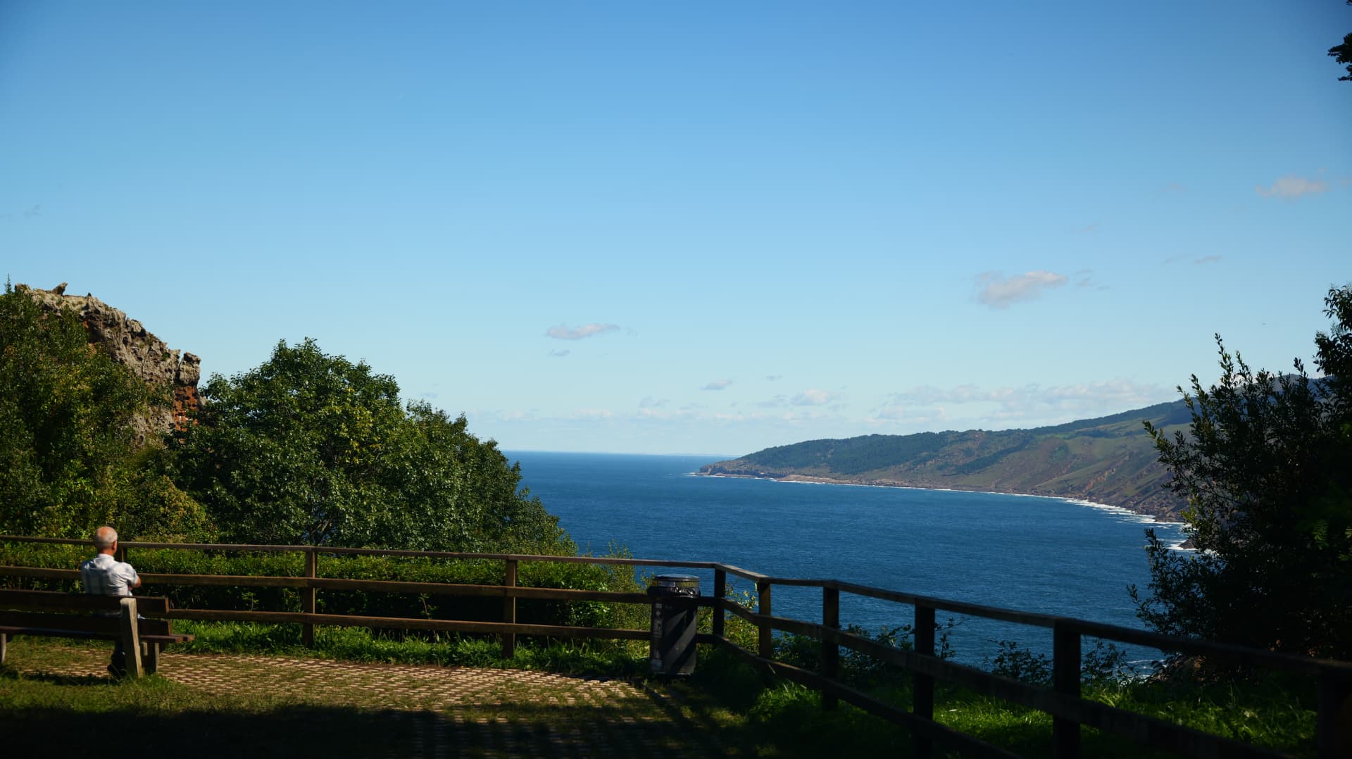 Man on a bench at a coastal viewpoint looking over the bay to distant green hills
