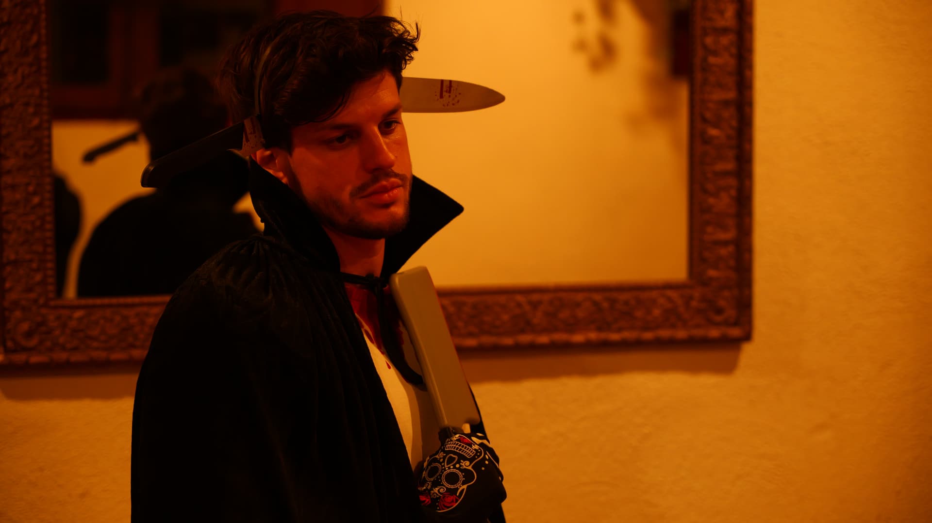 Man in a vampire cape and costume knives lit by warm lamplight beside a mirror