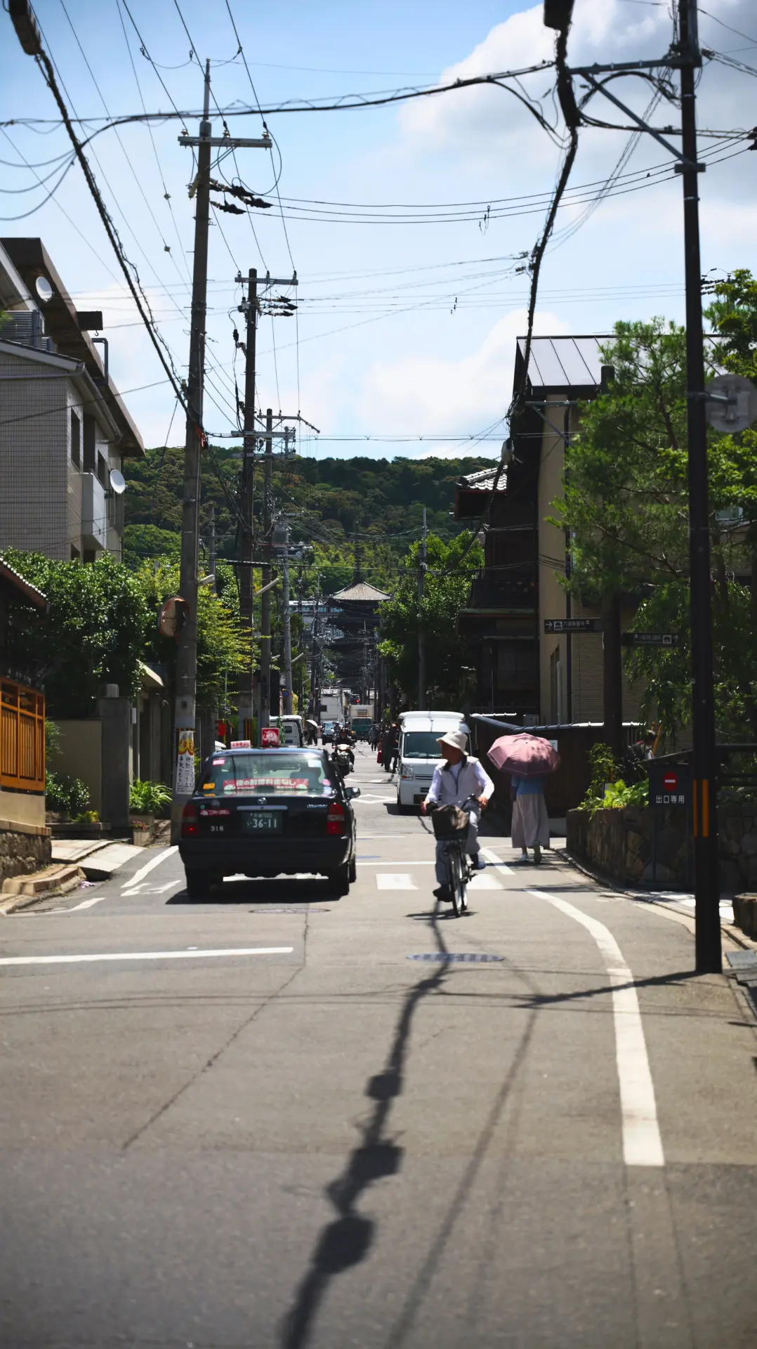 Taxi and cyclists on a sunlit Kyoto street with hills in the distance