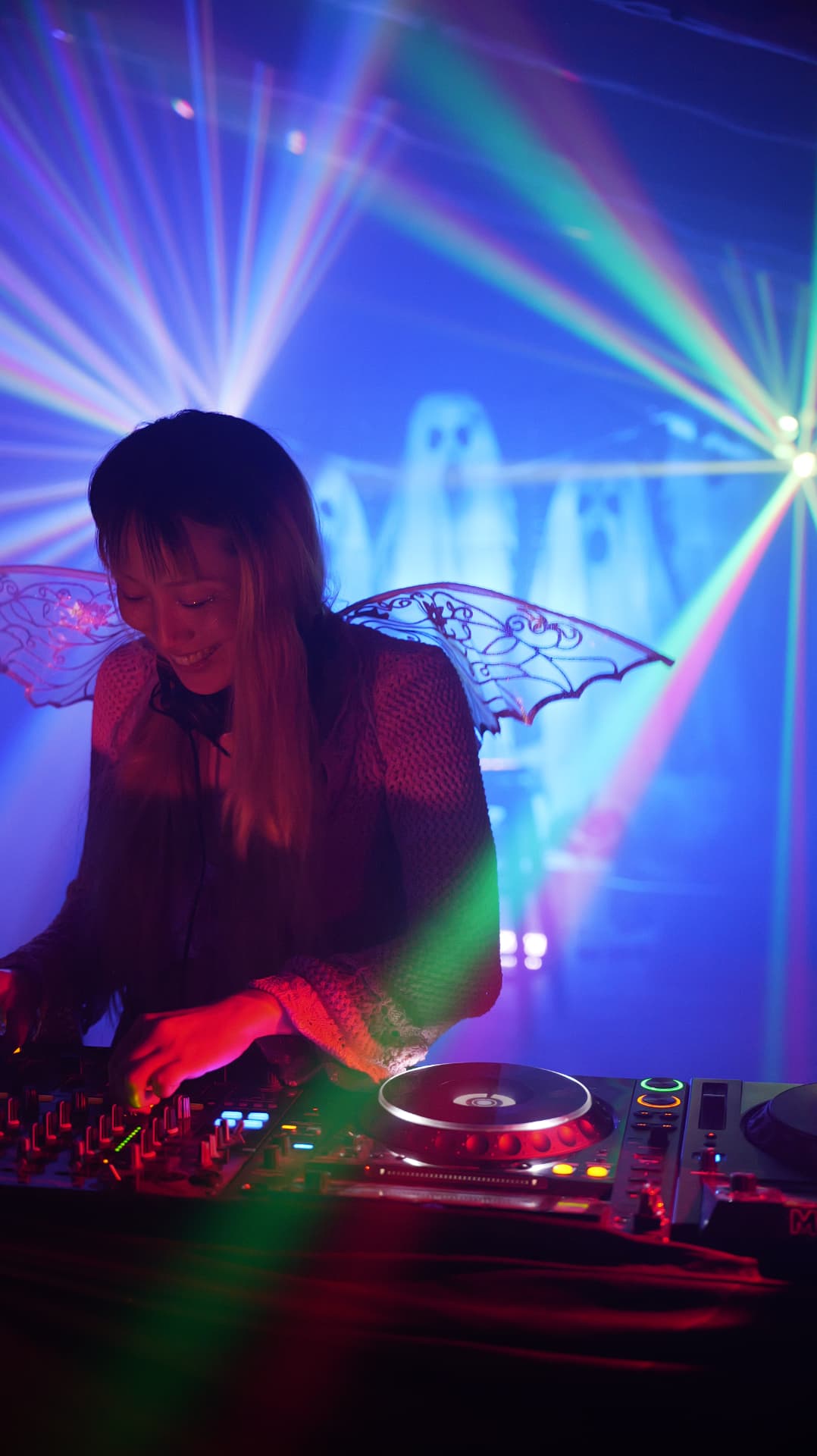 Smiling DJ in fairy wings at the decks under blue lasers and ghost projections