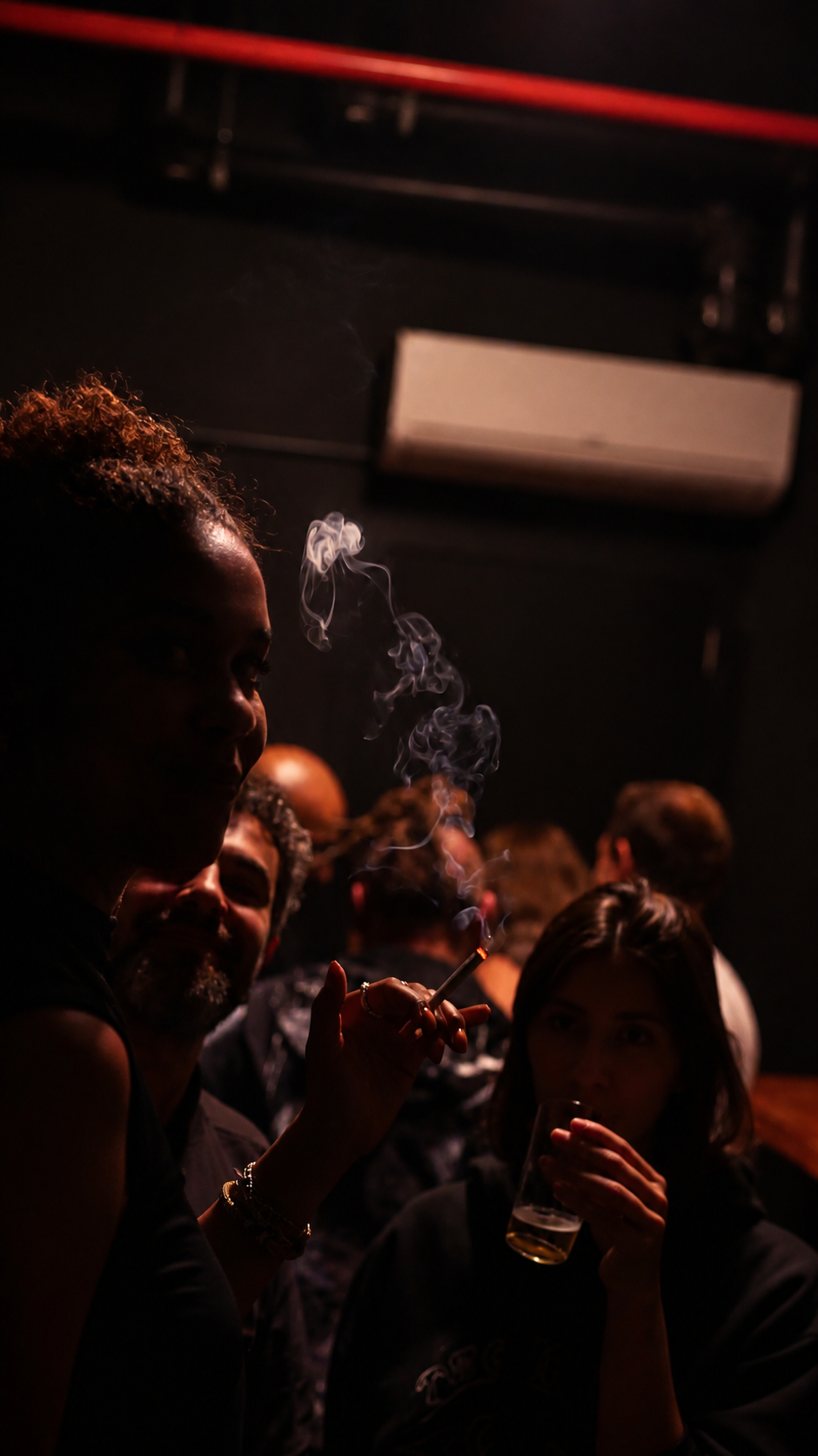 Woman smoking in a dark bar with faces lit by a faint red glow