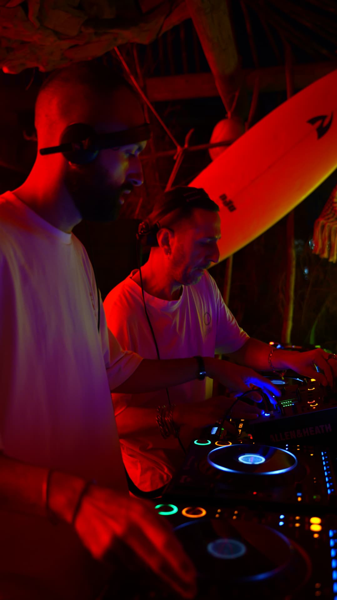 Two DJs mixing under red light in a beach bar hung with surfboards
