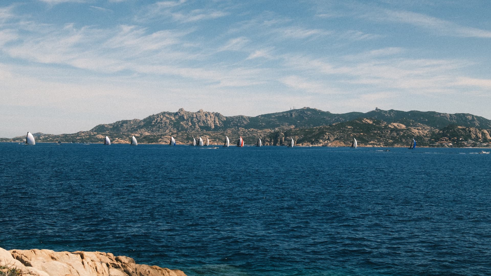 Sailing regatta crossing deep blue water below granite hills in Sardinia