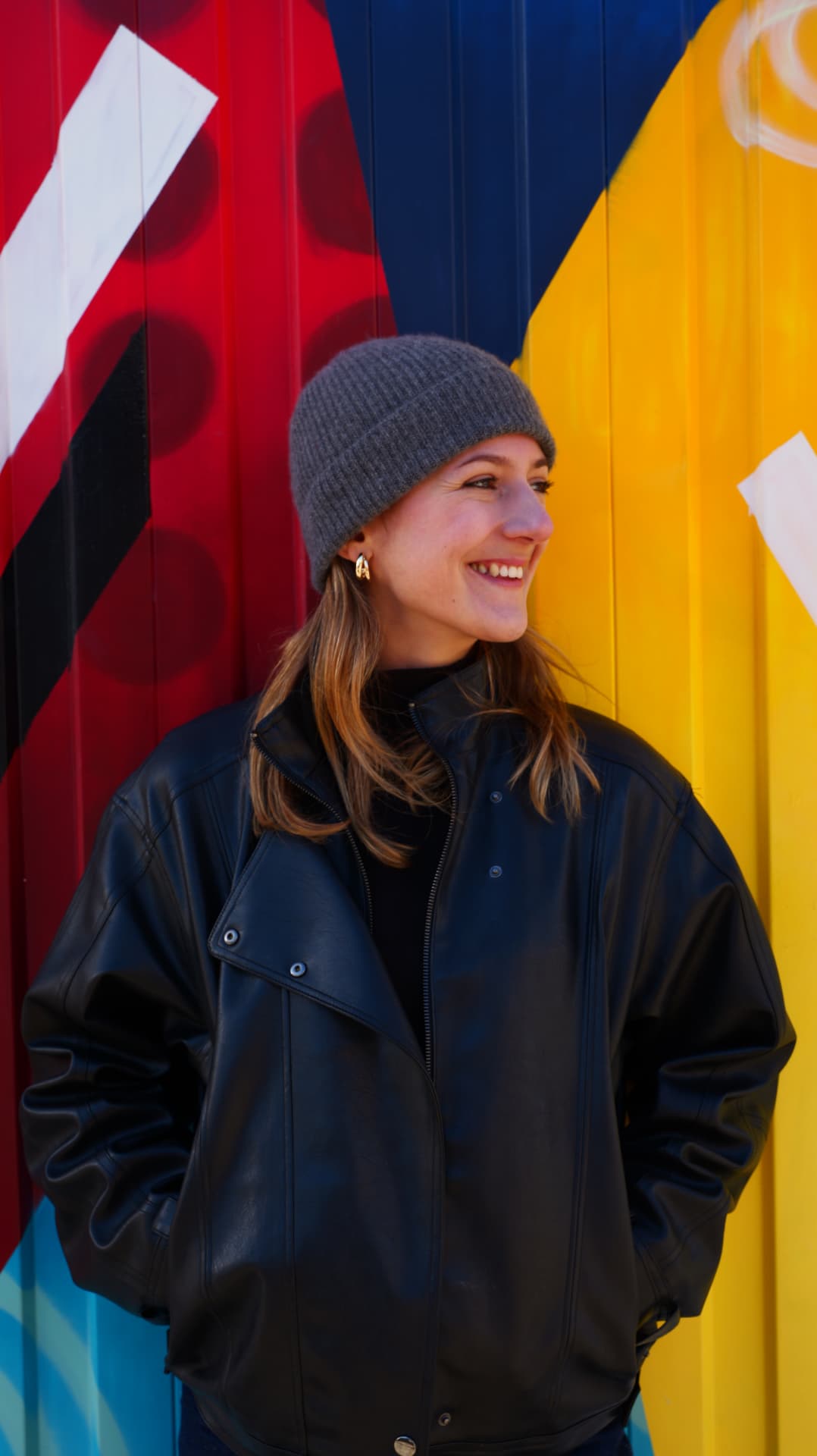 Smiling woman in a grey beanie and black leather jacket against red, yellow and blue stripes