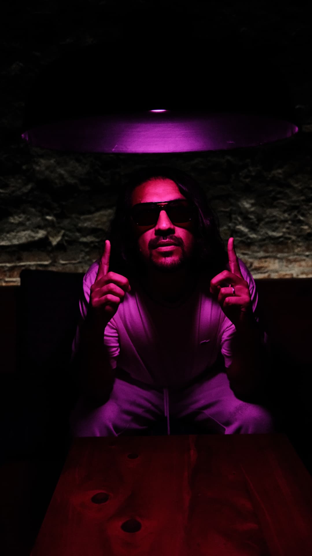 Portrait in red and purple club light, a man in sunglasses pointing up beneath a glowing lamp
